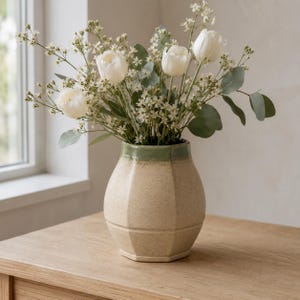 Puede incluir: Un jarrón de cerámica con cuerpo beige moteado y borde verde liso, que contiene un ramo de tulipanes blancos, pequeñas flores blancas en forma de estrella y hojas de eucalipto verdes. El jarrón tiene una forma redondeada y facetada.