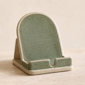 May include: A ceramic phone stand featuring an arched backrest with a glossy sage green glaze and a speckled cream-colored base. The stand has a sturdy, angled design with a small cutout in the front ledge to hold a mobile device securely in place.