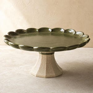 May include: A ceramic cake stand featuring a glossy, dark olive green circular top with a scalloped edge. The base is a sturdy, light beige, textured pedestal with a faceted, multi-sided design that supports the wide, flat serving surface.