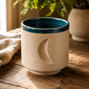 Könnte beinhalten: Ein zylindrischer Keramikbecher mit gesprenkelter beiger Außenseite und tiefem blaugrünem Inneren. Die Außenfläche weist eine eingeprägte Mondsichelform auf. Der Becher hat einen glatten, geraden Körper mit einem leicht vertieften Boden.