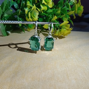 May include: A pair of silver drop earrings featuring rectangular, faceted green gemstones held by four-prong settings. The earrings are suspended from a thin silver metal rod, showcasing clear, vibrant green stones with polished silver hardware.