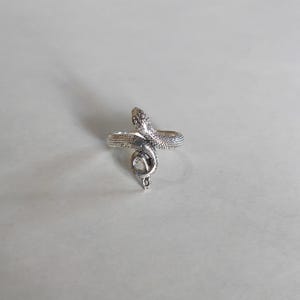 May include: A silver-toned metal ring shaped like a coiled snake. The band features a textured, scale-like pattern across the entire surface, with the snake's head positioned at the top and the tail curving downward to form the ring's circular band.