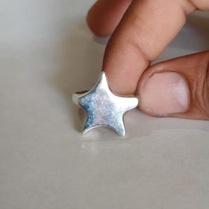 May include: A silver-toned ring featuring a prominent five-pointed star design on the front. The star has a smooth, reflective surface with subtle metallic highlights and a polished finish, attached to a simple, thin circular band.
