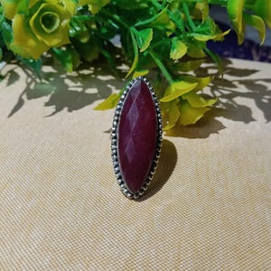 Op de afbeelding: Een zilveren ring met een grote, langwerpige, gefacetteerde donkerrode edelsteen in markiesvorm. De steen is gezet in een decoratieve zilveren zetting met een kralenrand, wat een textuurrijke omlijsting creëert rond het dieprode, geometrische oppervlak.