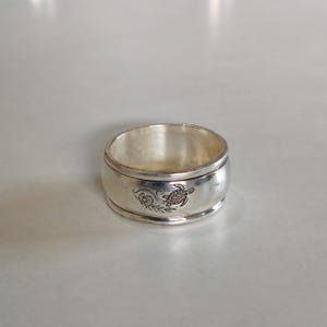 May include: A wide silver band ring featuring a polished metallic finish and a detailed engraved design of a sea turtle swimming alongside a small seashell and leafy sprig. The ring has a smooth, rounded outer surface with a clear, dark etched graphic.