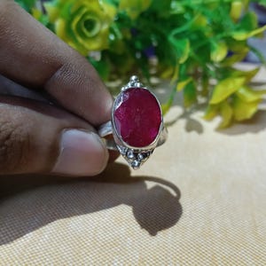 May include: A silver ring featuring a large, oval-shaped, faceted deep red gemstone set in a bezel. The metal band is decorated with small silver spheres at the top and bottom of the stone setting, creating a textured, metallic appearance.