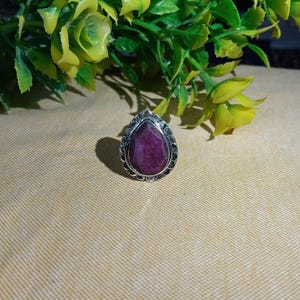 Può includere: Un anello in argento con una pietra preziosa a forma di pera color magenta intenso, incastonata in una cornice di metallo decorativa con bordo smerlato. La pietra ha una superficie opaca e testurizzata al centro della montatura in argento.