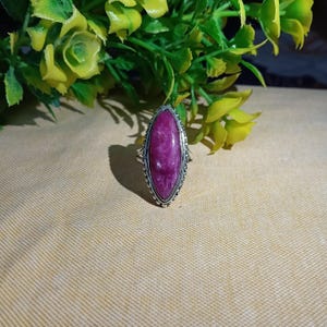 Pode incluir: Um anel em forma de marquise com uma pedra lisa de cor magenta profunda, engastada numa armação de metal prateado. O engaste apresenta um rebordo com contas que rodeia a gema oval, alongada e pontiaguda, com variações de cor naturais.