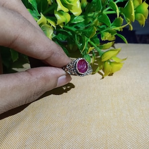 Op de afbeelding: Een zilveren ring met een centrale, ovaalvormige rode edelsteen. De metalen band is voorzien van ingewikkelde filigraan- en kraaldetails rondom de steen, wat zorgt voor een textuur. De ring wordt vastgehouden voor een achtergrond van groene bladeren.