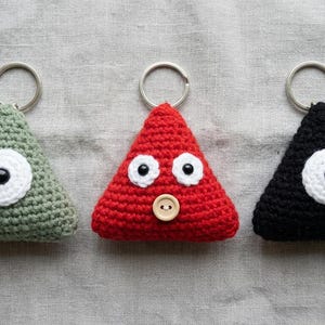 May include: Three triangular crochet keychains in sage green, bright red, and black. The green and black keychains feature a single central white eye with a black pupil. The red keychain has two white eyes with black pupils and a small beige button.