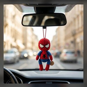 May include: A red and blue crocheted superhero figure hanging from a car rearview mirror. The figure features a round head with white eye patches, a spider emblem on the chest, and a small circular wooden tag attached to one arm.