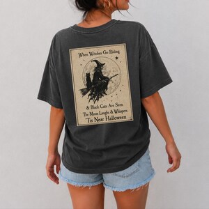 May include: A charcoal gray short-sleeve t-shirt featuring a rectangular graphic on the back. The print depicts a witch on a broomstick with a black cat against a full moon, with the text: When Witches Go Riding & Black Cats Are Seen The Moon Laughs & Whispers 'Tis Near Halloween.