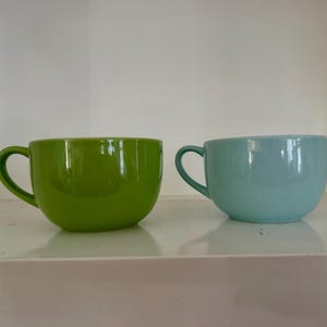May include: Two ceramic mugs with rounded bodies and curved handles. One mug is a vibrant lime green, while the other is a soft, pale blue. Both items feature a smooth, glossy finish and a wide, open top design, presented side by side.
