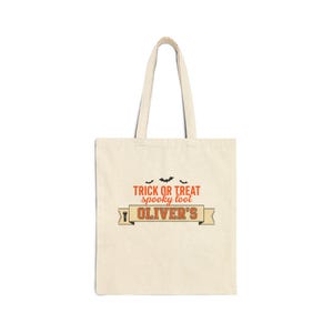 Peut inclure: Un sac fourre-tout en toile beige orné d'un motif avec trois chauves-souris noires et le texte orange Trick or Treat spooky loot. En dessous, une bannière contient le nom Oliver's en lettres orange, accompagné d'une petite icône de lampe torche.