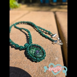 Op de afbeelding: Een ketting met een ovaalvormige steen met gevlekte groene en paarse patronen, gevat in een donkergroene macramé koordzetting. Het koord is geweven in een decoratief knoppatroon en bevat zilverkleurige metalen onderdelen bij de sluiting.