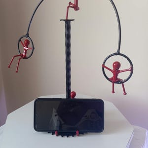 Könnte beinhalten: Ein Handyhalter aus Metall mit einem zentralen, gedrehten schwarzen Pfosten und einem horizontalen Bogen. Drei rote Strichmännchen sind an der Struktur befestigt, wobei zwei Figuren in schwarzen Ringen hängen und eine Figur den Bogen erklimmt.