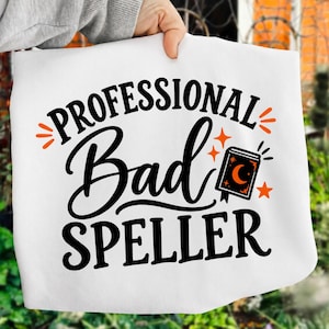 May include: A white t-shirt featuring the black text "PROFESSIONAL Bad SPELLER" in a mix of bold and script fonts. The design includes orange accents, specifically small stars, radiating lines, and a small book icon with a crescent moon.