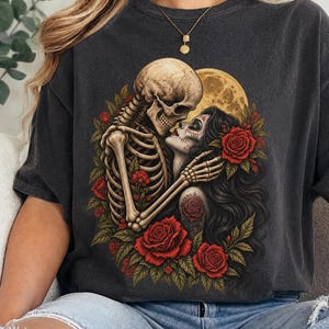 May include: A charcoal gray t-shirt featuring a graphic print of a skeleton embracing a person with sugar skull makeup. The design includes vibrant red roses, green leaves, and a large yellow full moon in the background, creating a detailed scene.