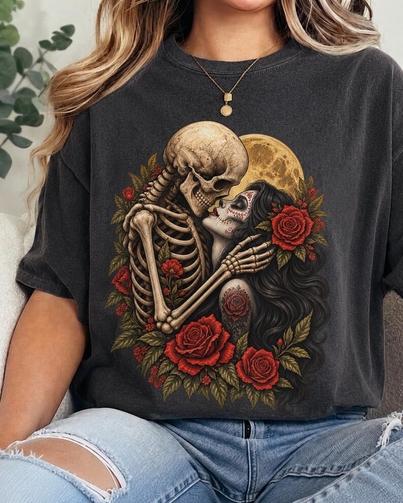 May include: A charcoal gray t-shirt featuring a graphic print of a skeleton embracing a person with sugar skull makeup. The design includes vibrant red roses, green leaves, and a large yellow full moon in the background, creating a detailed scene.
