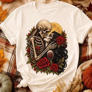 May include: A cream-colored short-sleeve t-shirt featuring a graphic print of a skeleton embracing a person with long dark hair and sugar skull makeup. The design includes red roses, green leaves, and a yellow full moon in the background.