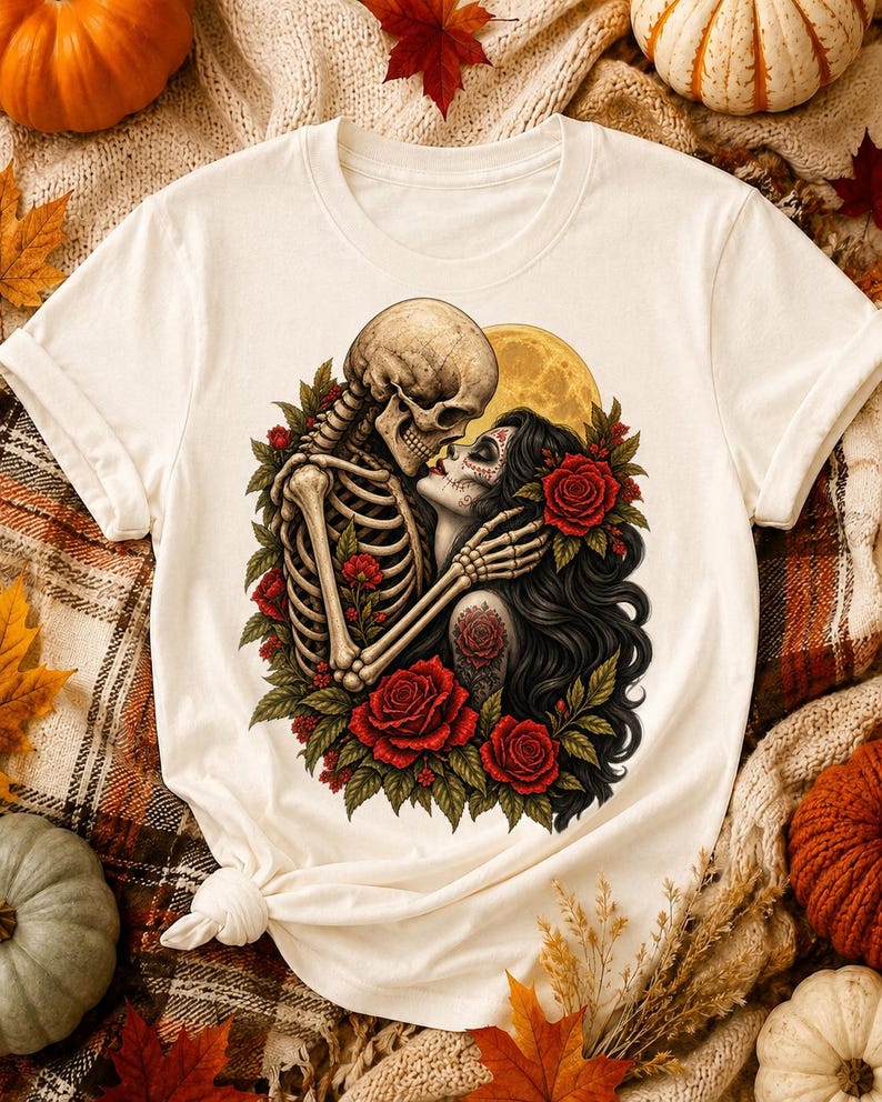 May include: A cream-colored short-sleeve t-shirt featuring a graphic print of a skeleton embracing a person with long dark hair and sugar skull makeup. The design includes red roses, green leaves, and a yellow full moon in the background.