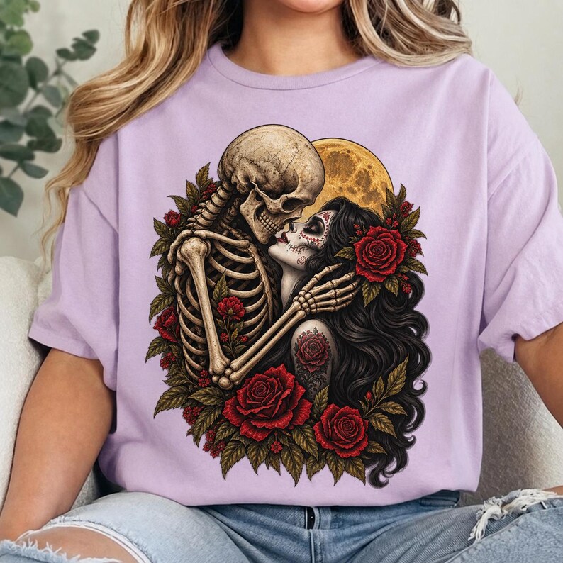 May include: A light purple t-shirt featuring a graphic print of a skeleton embracing a person with long dark hair and sugar skull makeup. The design includes vibrant red roses, green leaves, and a large yellow moon in the background.