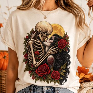 May include: A cream-colored t-shirt featuring a graphic print of a skeleton embracing a person with long black hair and sugar skull makeup. The design includes vibrant red roses, green leaves, and a large yellow moon in the background of the illustration.