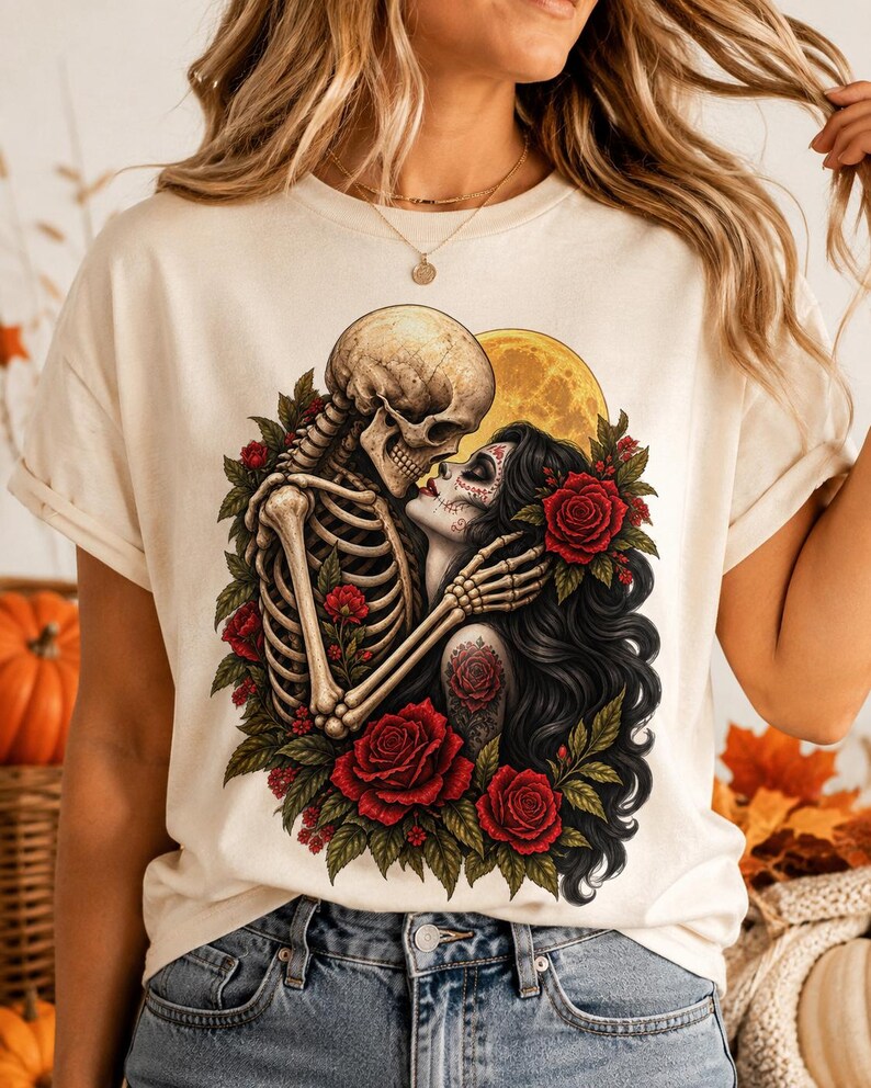 May include: A cream-colored t-shirt featuring a graphic print of a skeleton embracing a person with long black hair and sugar skull makeup. The design includes vibrant red roses, green leaves, and a large yellow moon in the background of the illustration.