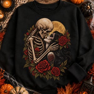 May include: A black crewneck sweatshirt featuring a graphic print of a skeleton embracing a person with sugar skull makeup. The design includes vibrant red roses, green leaves, and a large yellow full moon in the background, centered on the chest.
