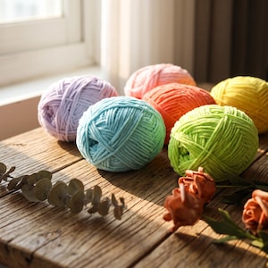 May include: Six round balls of thick, soft yarn in pastel and bright colors including lavender, light blue, lime green, orange, pink, and yellow. The yarn has a smooth, cord-like texture and is arranged in a cluster on a weathered wooden surface.