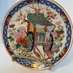 May include: A round porcelain decorative plate featuring a central illustration of a traditional ornate carriage surrounded by colorful chrysanthemums and cherry blossoms. The plate has a blue and white geometric patterned border with gold trim.