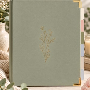 Könnte beinhalten: Ein salbeigrüner Ordner mit einer goldenen Blumen-Linienzeichnung auf dem Vorderdeckel. Der Ordner verfügt über goldene Metallecken und sechs farbige Registerkarten in Creme, Rosa, Olivgrün, Hellblau, Beige und Grau.