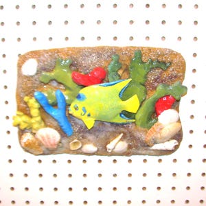 May include: A yellow and blue fish with a coral reef background. The fish is surrounded by coral, seaweed, and seashells. The background is a white pegboard.