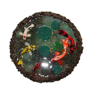 May include: A round resin art piece depicting a pond with green lily pads and colorful koi fish swimming. The pond is surrounded by a brown, textured border.