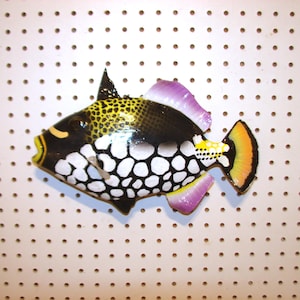 May include: A colorful, decorative fish figurine. The fish features a black body with white spots, yellow accents, and purple and orange fins. The fish is set against a white background with evenly spaced holes.