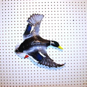 K&ouml;nnte beinhalten: Eine dekorative Stockente im Flug, mit schwarzem K&ouml;rper, wei&szlig;en Akzenten und blauen Fl&uuml;geldetaills. Die Ente hat einen gelben Schnabel und rote F&uuml;&szlig;e. Das Bild ist vor einem wei&szlig;en Hintergrund mit gleichm&auml;&szlig;ig verteilten braunen Punkten.