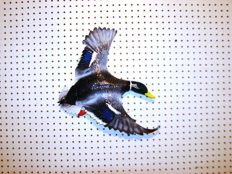 K&ouml;nnte beinhalten: Eine dekorative Stockente im Flug, mit schwarzem K&ouml;rper, wei&szlig;en Akzenten und blauen Fl&uuml;geldetaills. Die Ente hat einen gelben Schnabel und rote F&uuml;&szlig;e. Das Bild ist vor einem wei&szlig;en Hintergrund mit gleichm&auml;&szlig;ig verteilten braunen Punkten.