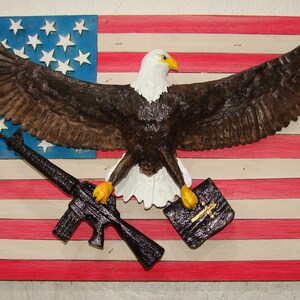 May include: A carved wooden bald eagle with outstretched wings holding a rifle and a book with a cross on it. The eagle is positioned in front of a painted American flag.