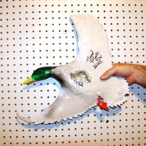 K&ouml;nnte beinhalten: Eine handbemalte, wei&szlig;e Entenskulptur mit gr&uuml;nem Kopf, gelbem Schnabel und roten Akzenten. Die Skulptur hat eine strukturierte Oberfl&auml;che und wird vor einer wei&szlig;en Lochplatte gehalten. Der Fl&uuml;gel der Ente ist ausgestreckt und tr&auml;gt den Text "Brad Martin 2025" und die Nummer "552".