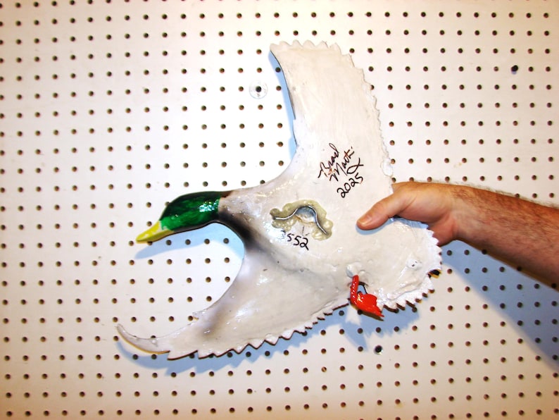 K&ouml;nnte beinhalten: Eine handbemalte, wei&szlig;e Entenskulptur mit gr&uuml;nem Kopf, gelbem Schnabel und roten Akzenten. Die Skulptur hat eine strukturierte Oberfl&auml;che und wird vor einer wei&szlig;en Lochplatte gehalten. Der Fl&uuml;gel der Ente ist ausgestreckt und tr&auml;gt den Text "Brad Martin 2025" und die Nummer "552".
