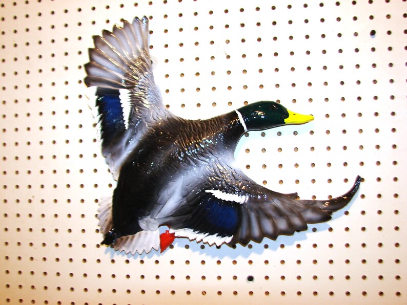 K&ouml;nnte beinhalten: Detaillierte, dreidimensionale Entenskulptur im Flug. Die Ente hat eine gl&auml;nzende Oberfl&auml;che mit einem dunklen K&ouml;rper, wei&szlig;en und blauen Fl&uuml;gelakzenten und einem gelben Schnabel. Die Skulptur ist auf einer wei&szlig;en Lochplatte montiert.