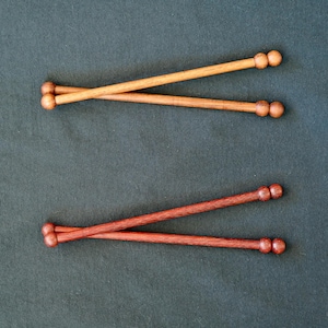 May include: Two pairs of wooden knitting needles arranged in an X shape. The top pair is light brown, and the bottom pair is a darker reddish-brown. Each needle features a smooth, cylindrical shaft and a small, rounded knob at the end.