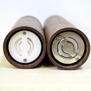 May include: Two wooden salt and pepper grinders with a dark brown finish. One grinder has a white plastic mechanism, while the other features a stainless steel grinding mechanism. The grinders are cylindrical and designed for kitchen use.