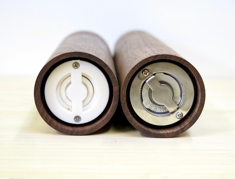 May include: Two wooden salt and pepper grinders with a dark brown finish. One grinder has a white plastic mechanism, while the other features a stainless steel grinding mechanism. The grinders are cylindrical and designed for kitchen use.