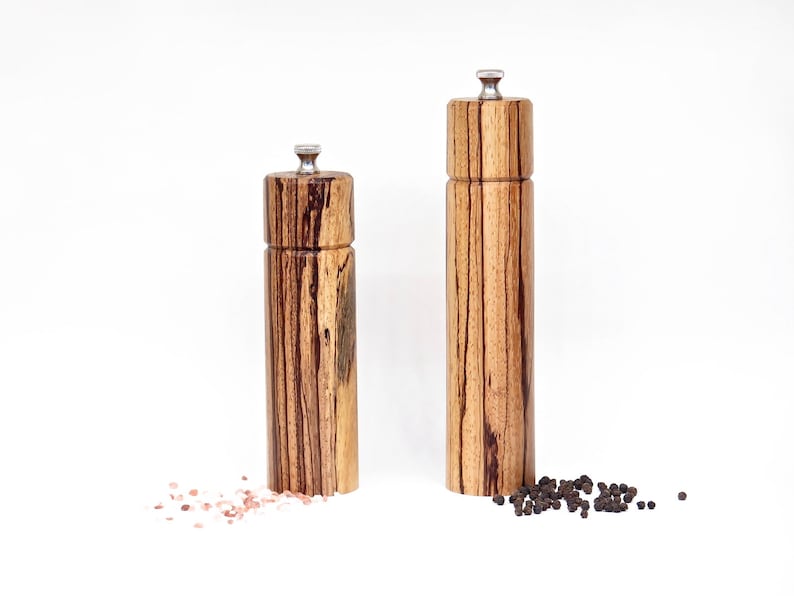 K&ouml;nnte beinhalten: Zwei Salz- und Pfefferm&uuml;hlen aus Holz mit einem gestreiften Holzmaserungsmuster. Die M&uuml;hlen haben einen silbernen Deckel und stehen auf einer wei&szlig;en Oberfl&auml;che. Rosa Himalaya-Salz ist um die kleinere M&uuml;hle herum verstreut und schwarze Pfefferk&ouml;rner sind um die gr&ouml;&szlig;ere M&uuml;hle herum verstreut.