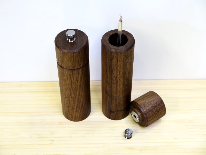May include: A set of three wooden pepper mills with a dark brown finish. One mill is assembled, another is partially disassembled, and the third is open, revealing the grinding mechanism. The mills have a cylindrical shape with a silver top.