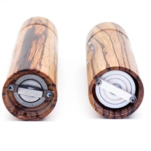 K&ouml;nnte beinhalten: Zwei Salz- und Pfefferm&uuml;hlen aus Holz mit einem Edelstahlmechanismus. Die M&uuml;hlen sind aus Holz mit einem dunkelbraunen und hellbraunen Streifenmuster. Die Unterseite einer M&uuml;hle zeigt die Worte "Stainless Steel" und "Made in U.S.A.". Die andere M&uuml;hle zeigt den inneren Mechanismus mit einem wei&szlig;en Keramikmahlwerk.