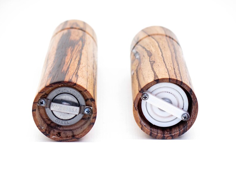 K&ouml;nnte beinhalten: Zwei Salz- und Pfefferm&uuml;hlen aus Holz mit einem Edelstahlmechanismus. Die M&uuml;hlen sind aus Holz mit einem dunkelbraunen und hellbraunen Streifenmuster. Die Unterseite einer M&uuml;hle zeigt die Worte "Stainless Steel" und "Made in U.S.A.". Die andere M&uuml;hle zeigt den inneren Mechanismus mit einem wei&szlig;en Keramikmahlwerk.