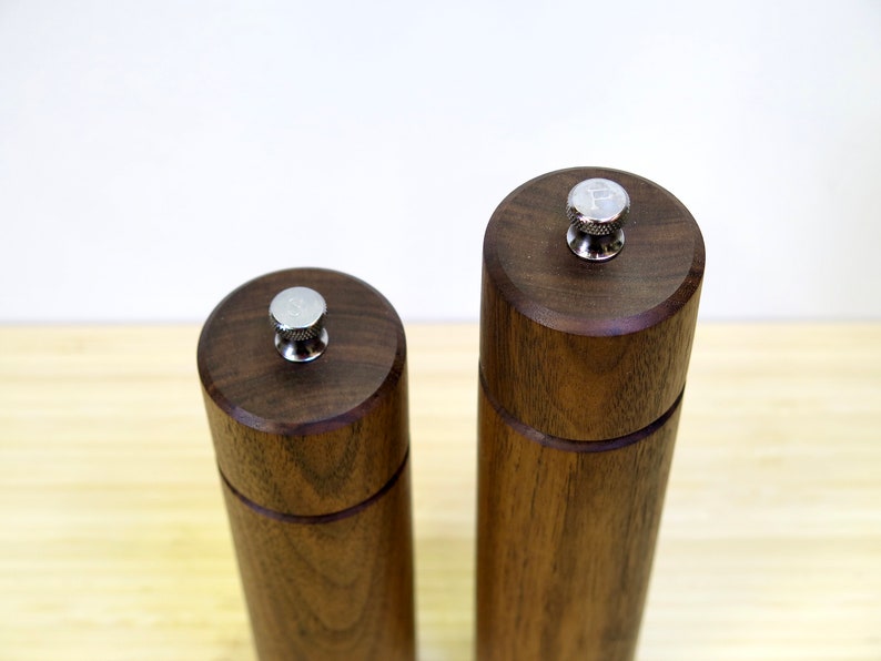 May include: Two wooden salt and pepper grinders. The grinders are made of dark brown wood with a cylindrical shape. Each grinder has a silver-colored top with a letter on it, one with an "S" and the other with a "P".