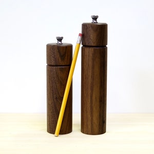 May include: Two wooden salt and pepper grinders with a pencil for scale. The grinders are made of dark brown wood with a cylindrical shape and a silver-colored top. The pencil is yellow with a pink eraser.